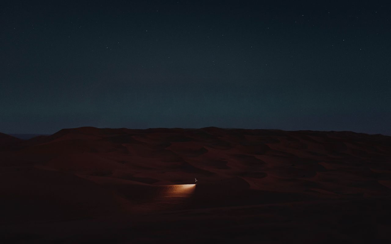 A lone explorer illuminates a path through dark desert dunes beneath a star-filled sky.